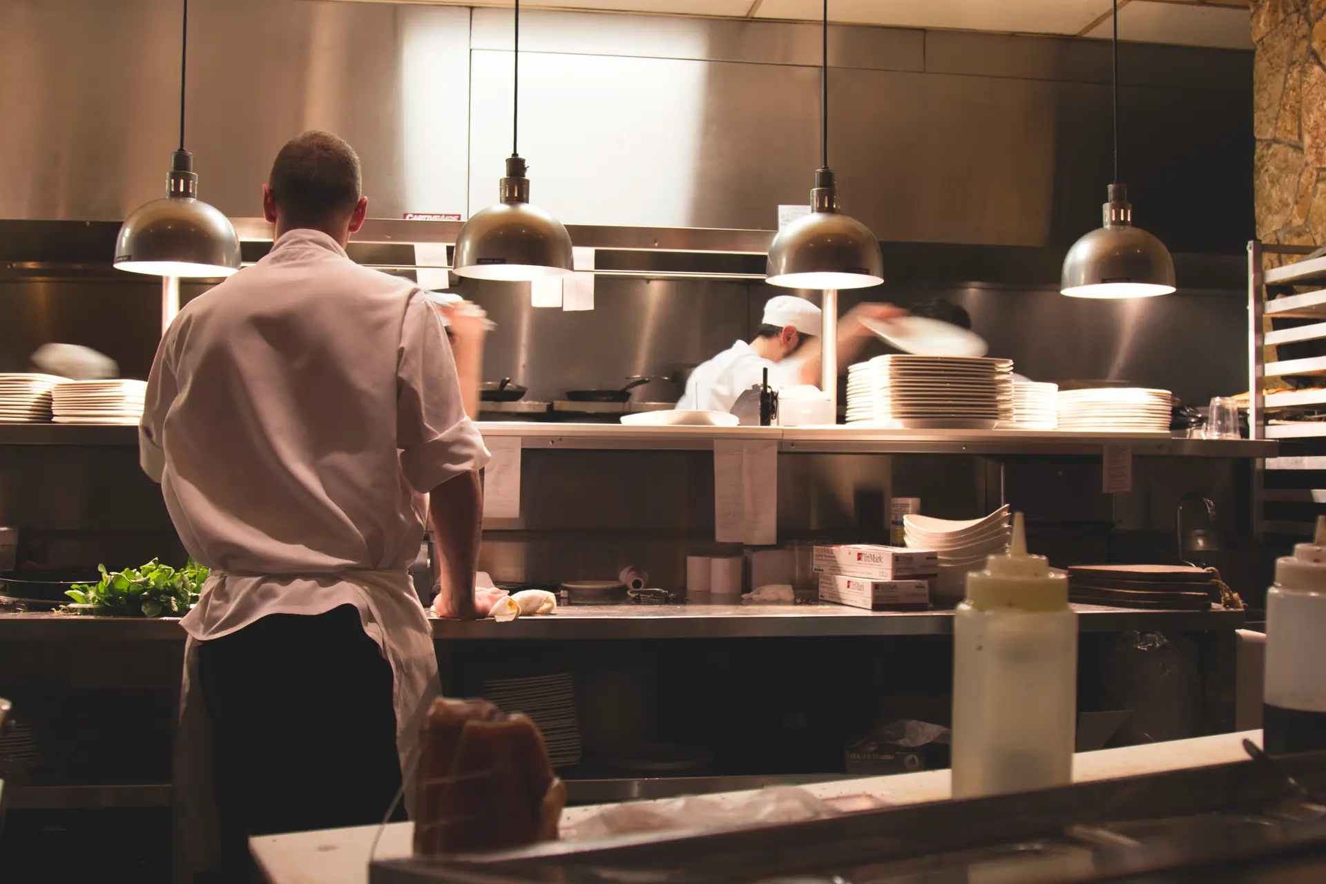 Le passe sous ses quatre lampes, un cuisinier de dos, les assiettes empilées